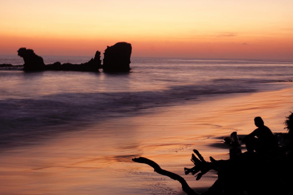 A red and orange sky at sunset is reflected on the calm waters and fine sand