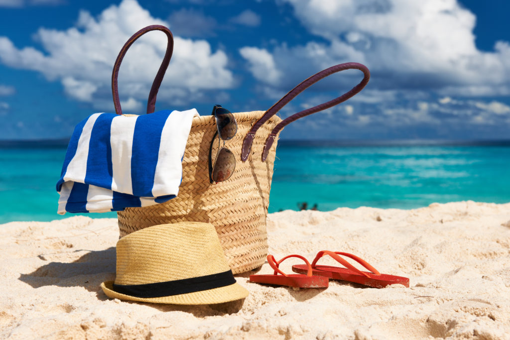 Beach bag with a beach towel and sunglasses spilling out of it, on the sand with a sun hat and flip flops on the ground